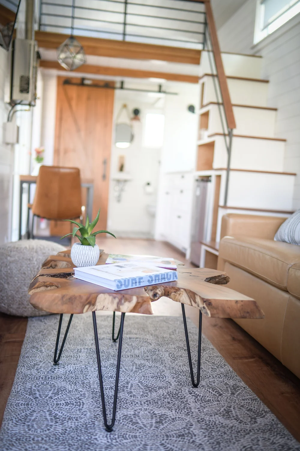 Living room with a wooden coffee table, a tan leather couch, a potted plant, magazine, and books on the table. In the background, stairs, a door, and a small white bathroom are visible.