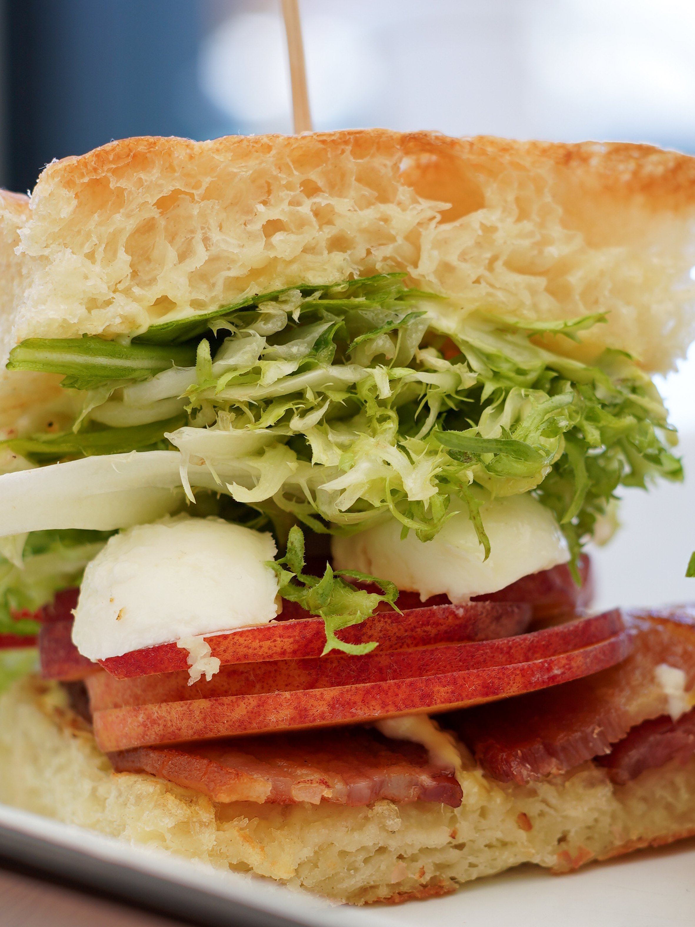 Close-up of a layered sandwich with bacon, sliced tomatoes, mozzarella cheese, shredded lettuce, and a fluffy bread bun.