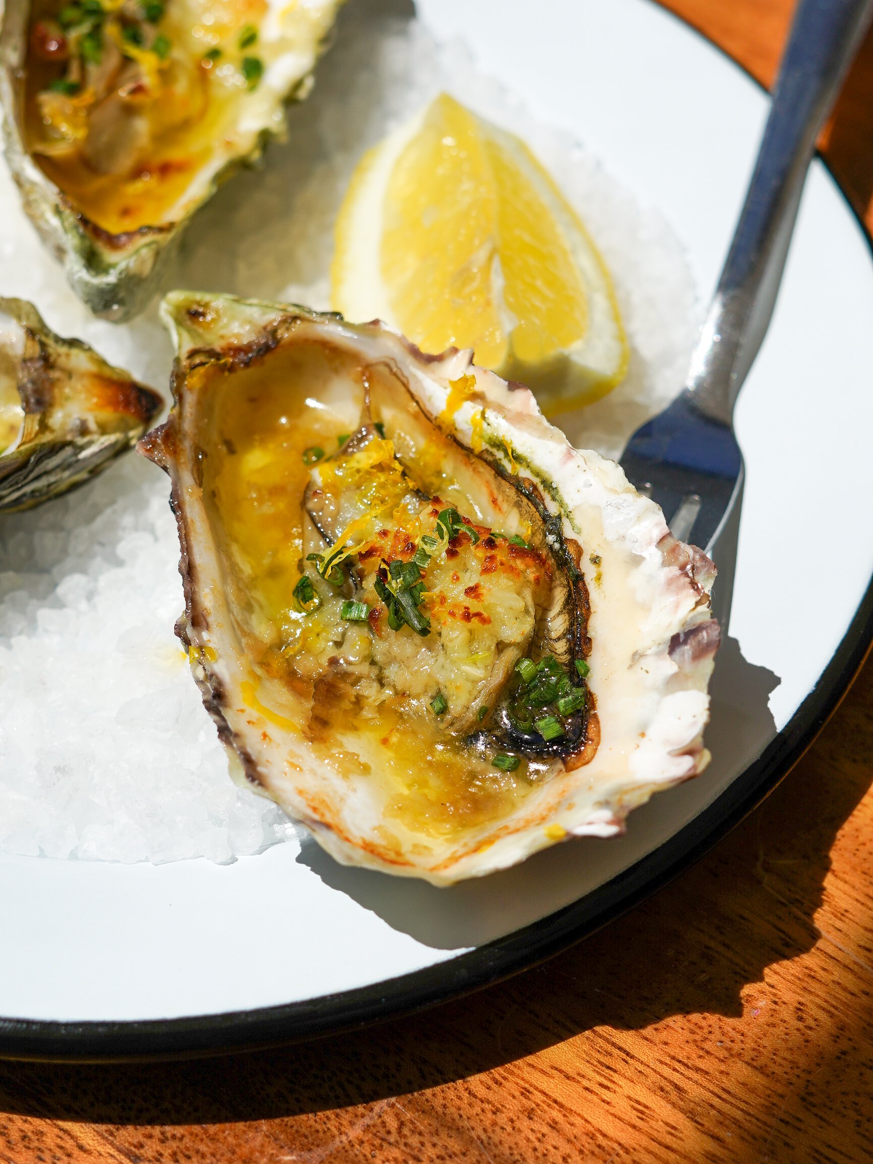 A close-up of oysters on a bed of ice with lemon wedges, garnished with green herbs and spice.