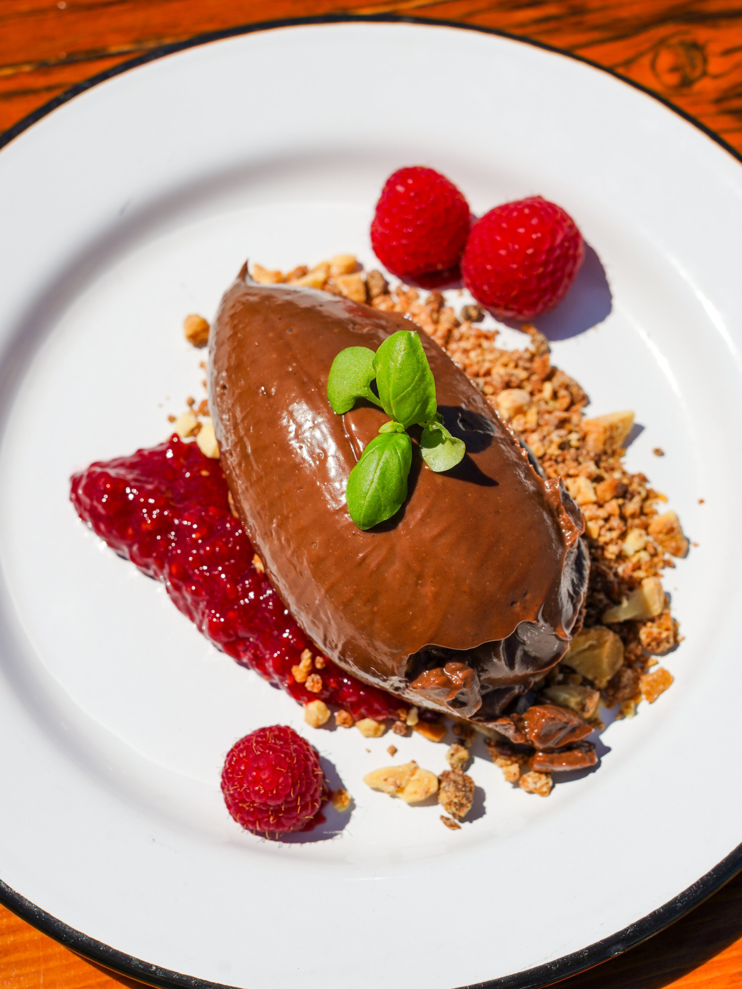 A dessert on a white plate with raspberries, chopped nuts, a chocolate-covered item, and a small green herb garnish.