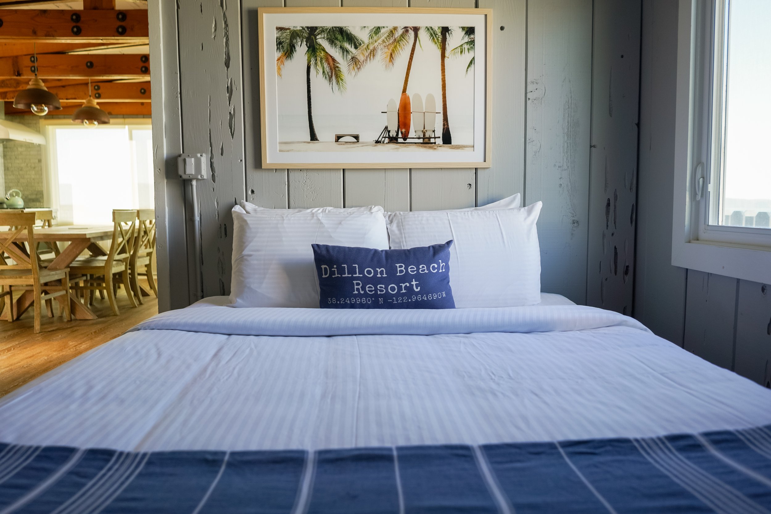 A bright bedroom at ブライトリビエラ with a white-striped duvet, a navy blue accent pillow, and tropical palm tree wall art. In the background, a warm wooden dining area is visible through an open doorway.