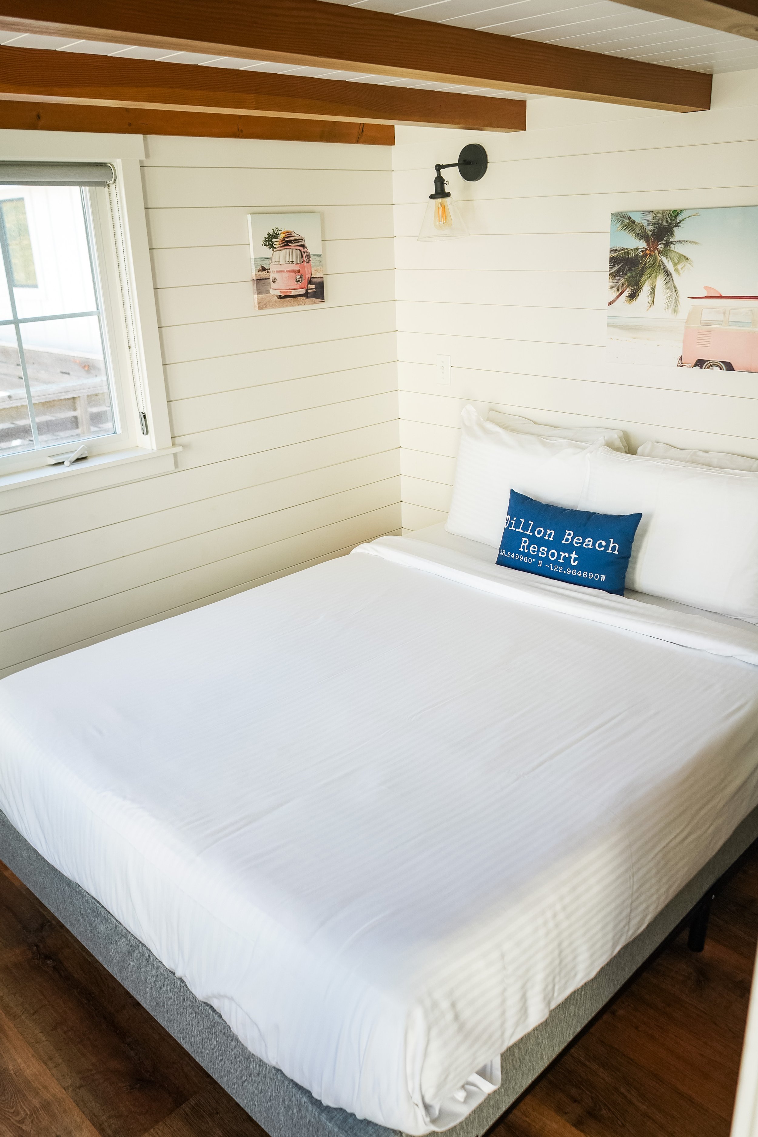 A neatly made white bed with a blue pillow that says 'ブライトリビエラ' in a room with shiplap walls, two wall-mounted light fixtures, and artwork featuring a vintage pink van, surfboard, palm trees, and a beach scene.