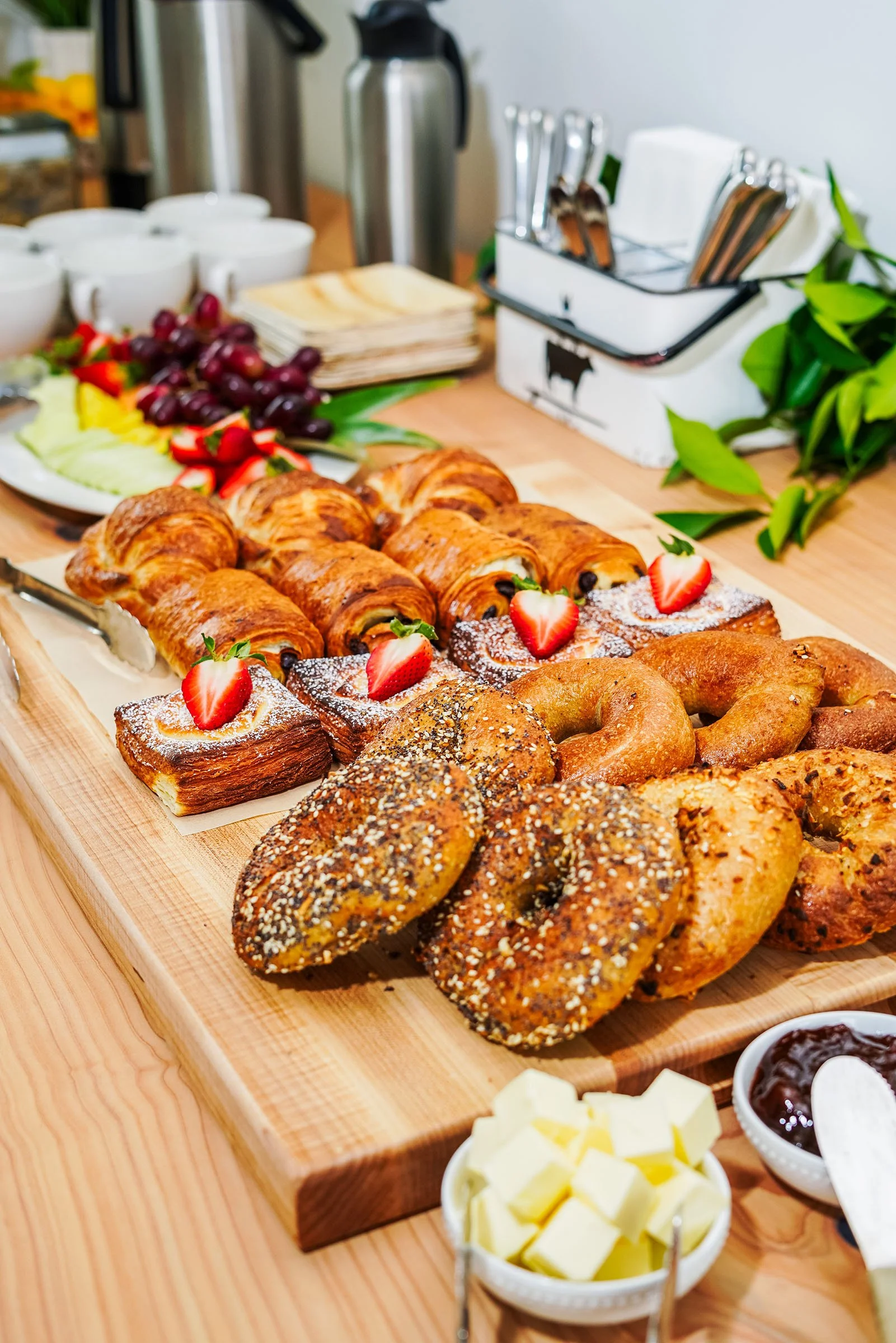 Variety of baked pastries, including croissants, and sliced strawberries on a wooden table with bowls of butter, jam, and cheese, and a fruit platter with grapes, strawberries, and sliced melons.