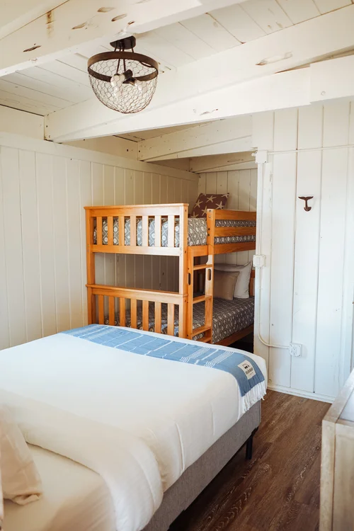 A family-friendly bedroom with white wood-paneled walls and exposed ceiling beams. The room features a queen bed with white linens and a blue throw, and a wooden twin-over-twin bunk bed tucked into an alcove.