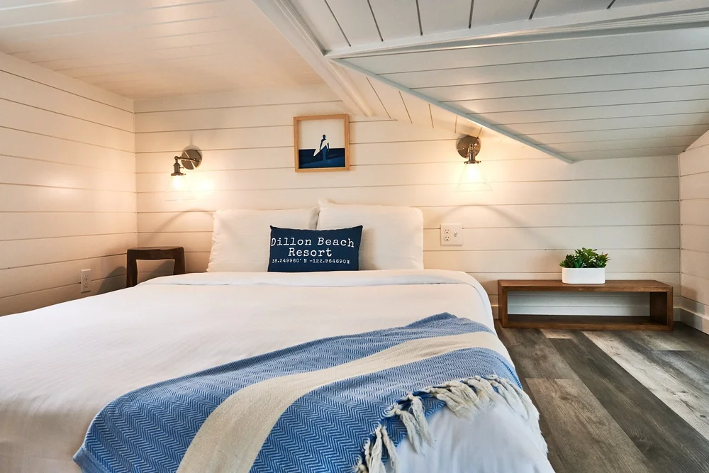 View from inside a modern beach cabin looking out through glass doors to a private stone patio with a picnic table and fire pit. The interior features a white farmhouse sink and a dining island with leather stools.