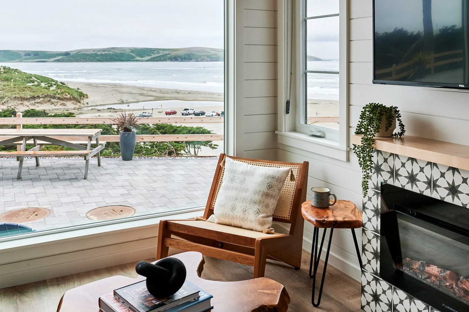 Panoramic view of the beach and ocean from a modern ブライトリビエラ COHO cabin. Inside, a mid-century style cane chair with a patterned pillow sits on a live-edge coffee table, next to an electric fireplace with patterned tile and a live-edge man