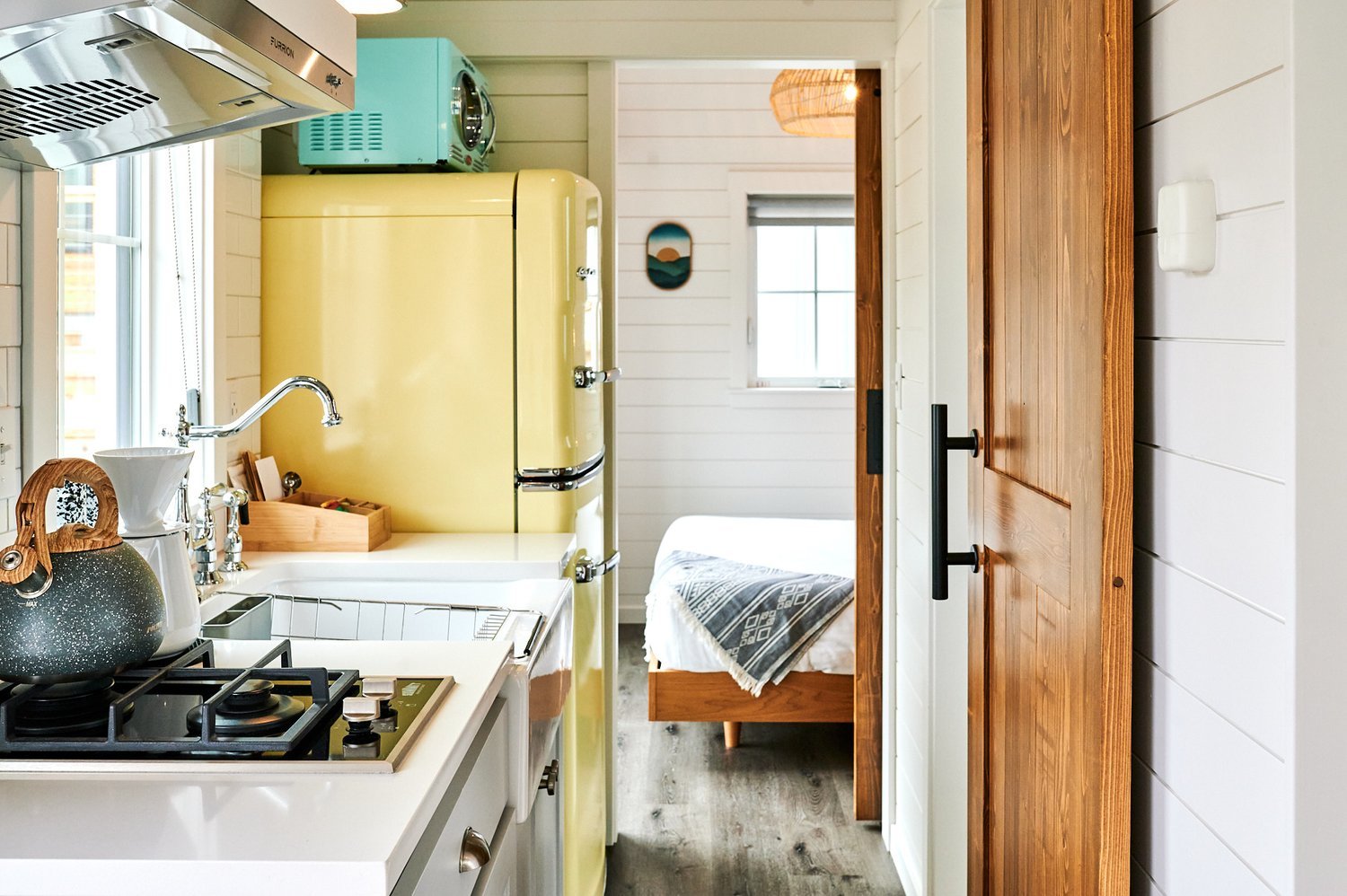 A view from the Sandpiper cabin kitchen through an open doorway to the bedroom. It shows a modern two-burner stove, a vintage-style yellow refrigerator, a turquoise microwave, and a glimpse of the shiplap-walled bedroom beyond.