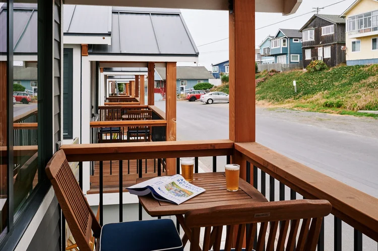 The private wooden deck of a Sandpiper cabin looking out at a row of similar tiny homes. A small slatted wood bistro table with two glasses of beer and an open book sits next to matching wooden chairs.