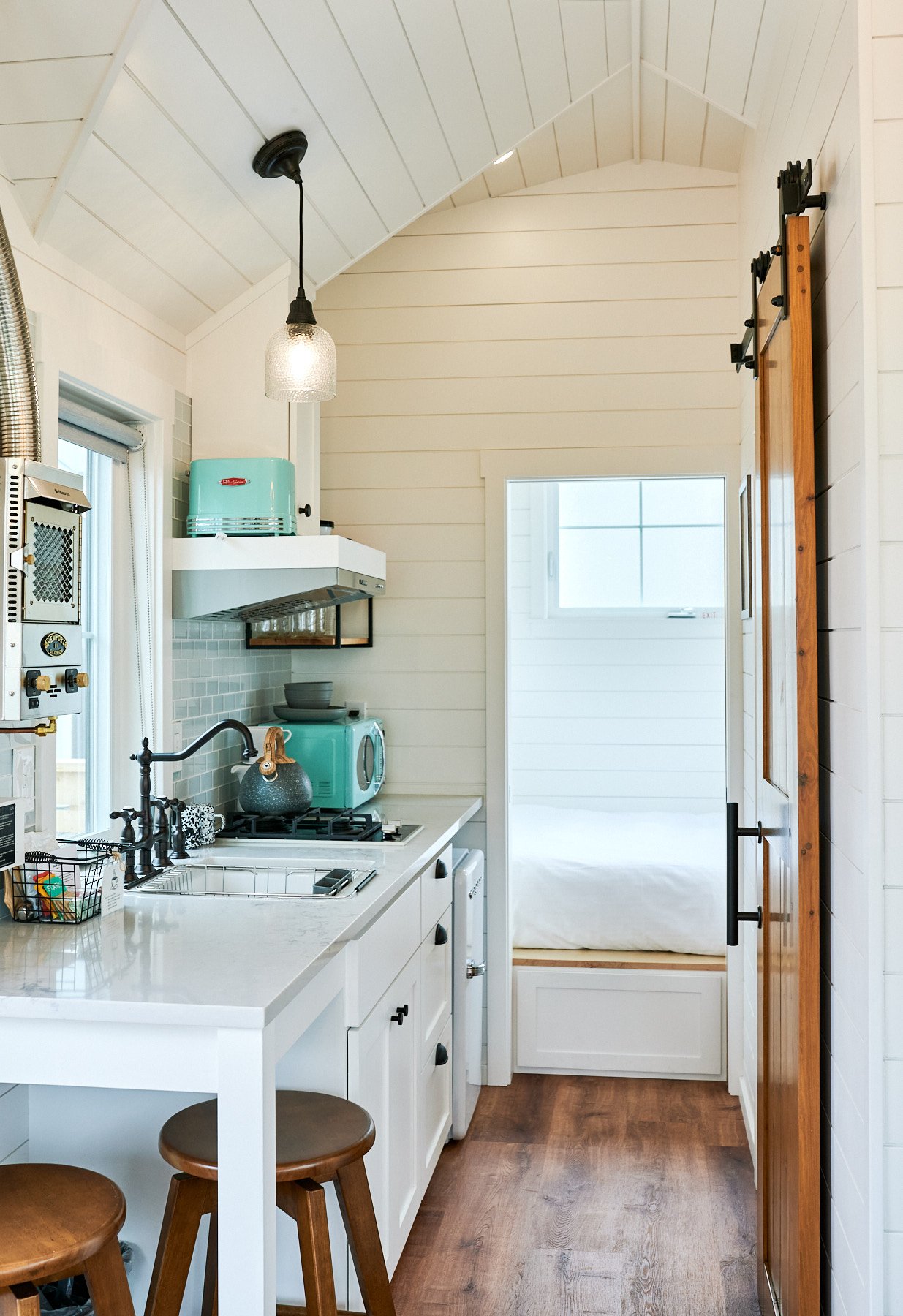 A small, modern kitchen with white cabinets, a white countertop, a small sink, a gas stove, and a microwave. There are two wooden stools at a white counter, and a hanging light fixture. The kitchen has white shiplap walls and ceiling, and a sliding b