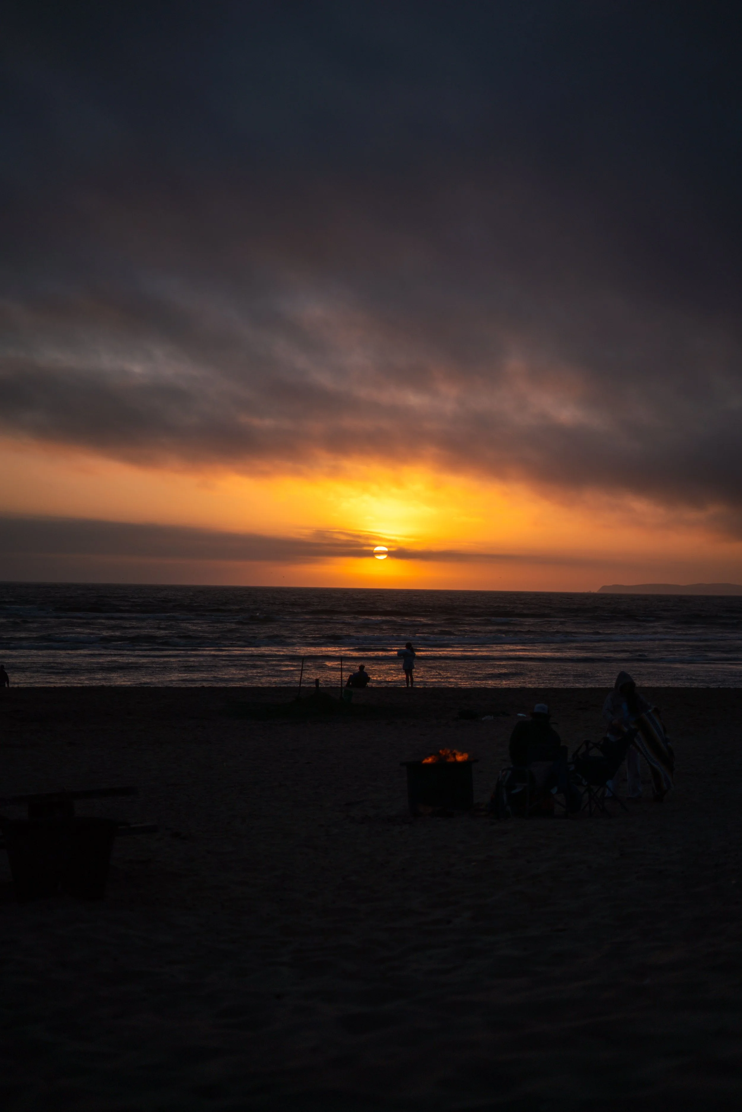 A sunset over the ocean with dark clouds in the sky and a few people sitting and standing on the beach, some near a small fire.