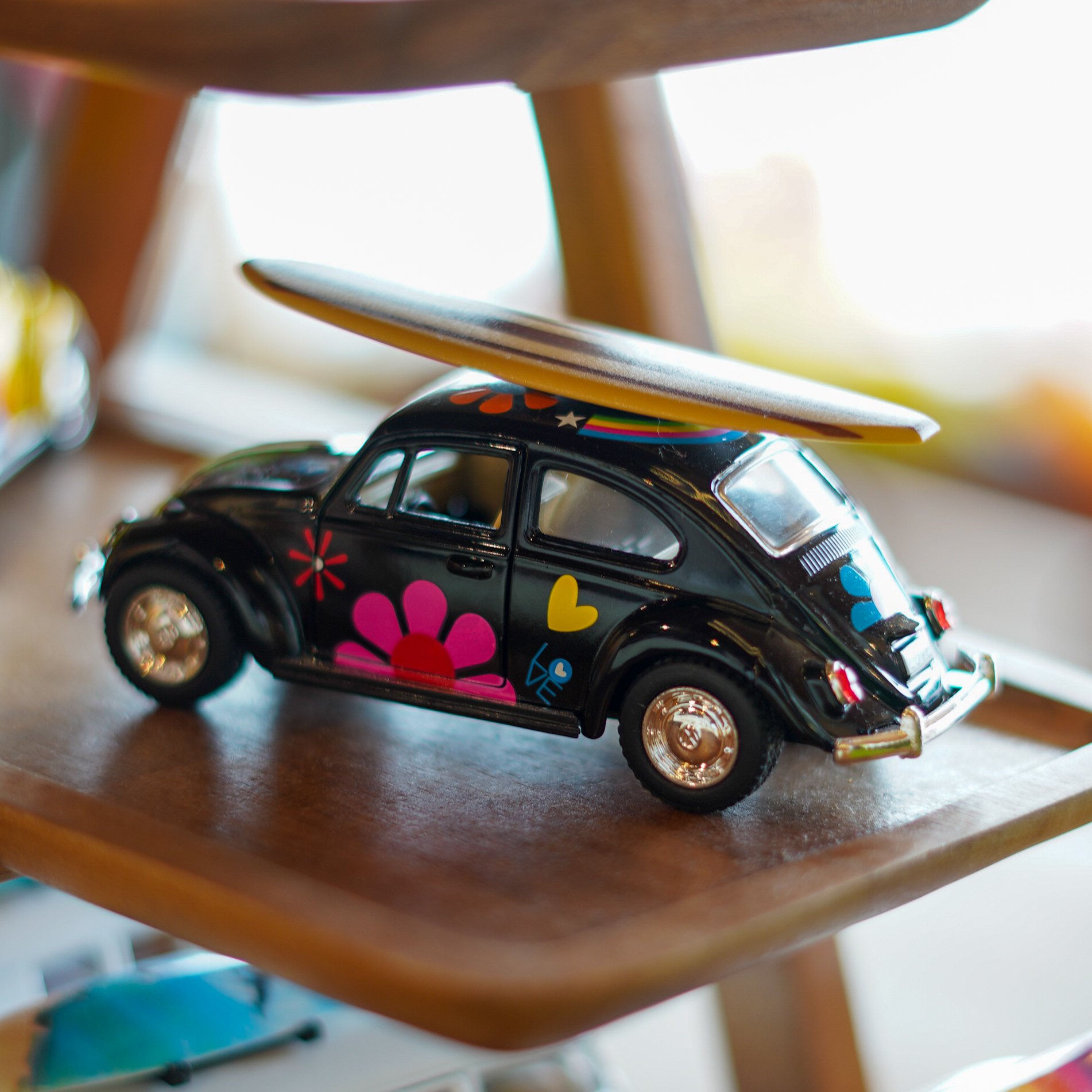 Close-up of a toy car with colorful flower and heart decals, stacked beneath a wooden furniture piece, with a skateboard and other objects in the background.