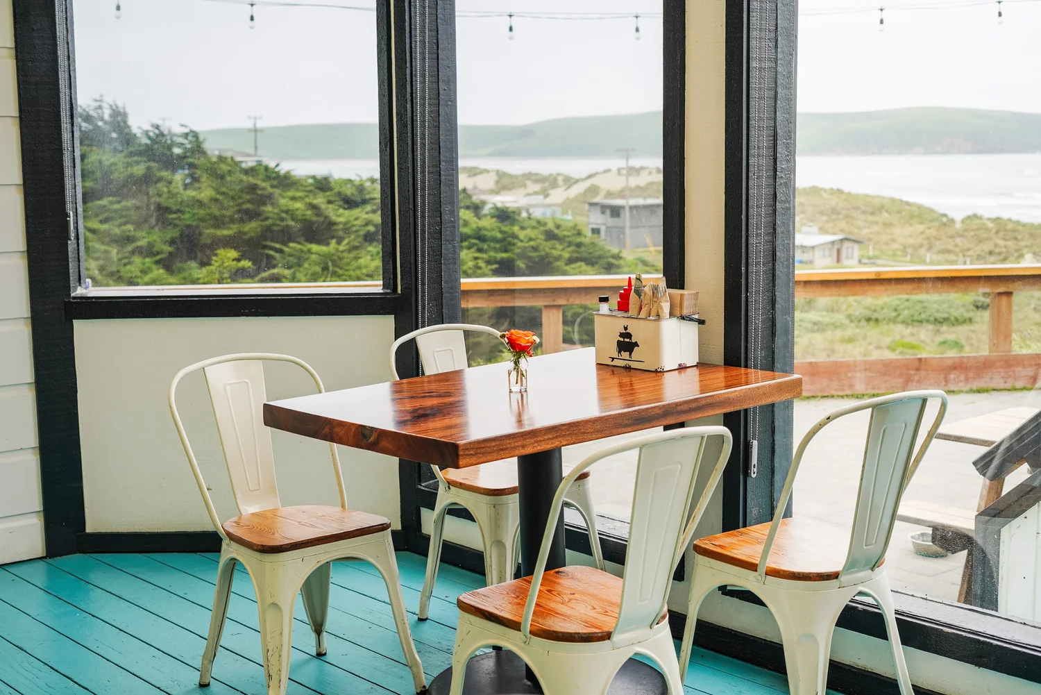 Interior view of a dining area with a wooden table, white chairs, and a view of greenery and hills outside through open windows.