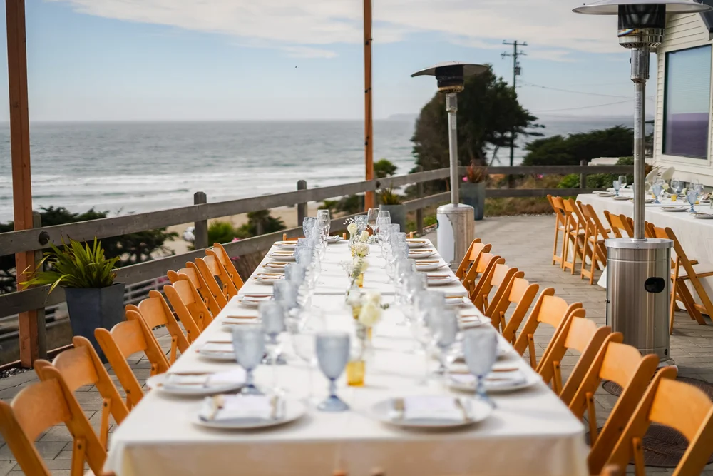 A long outdoor dining table set with white tablecloths, glassware, and floral centerpieces overlooking the ocean with a beach and sky in the background.