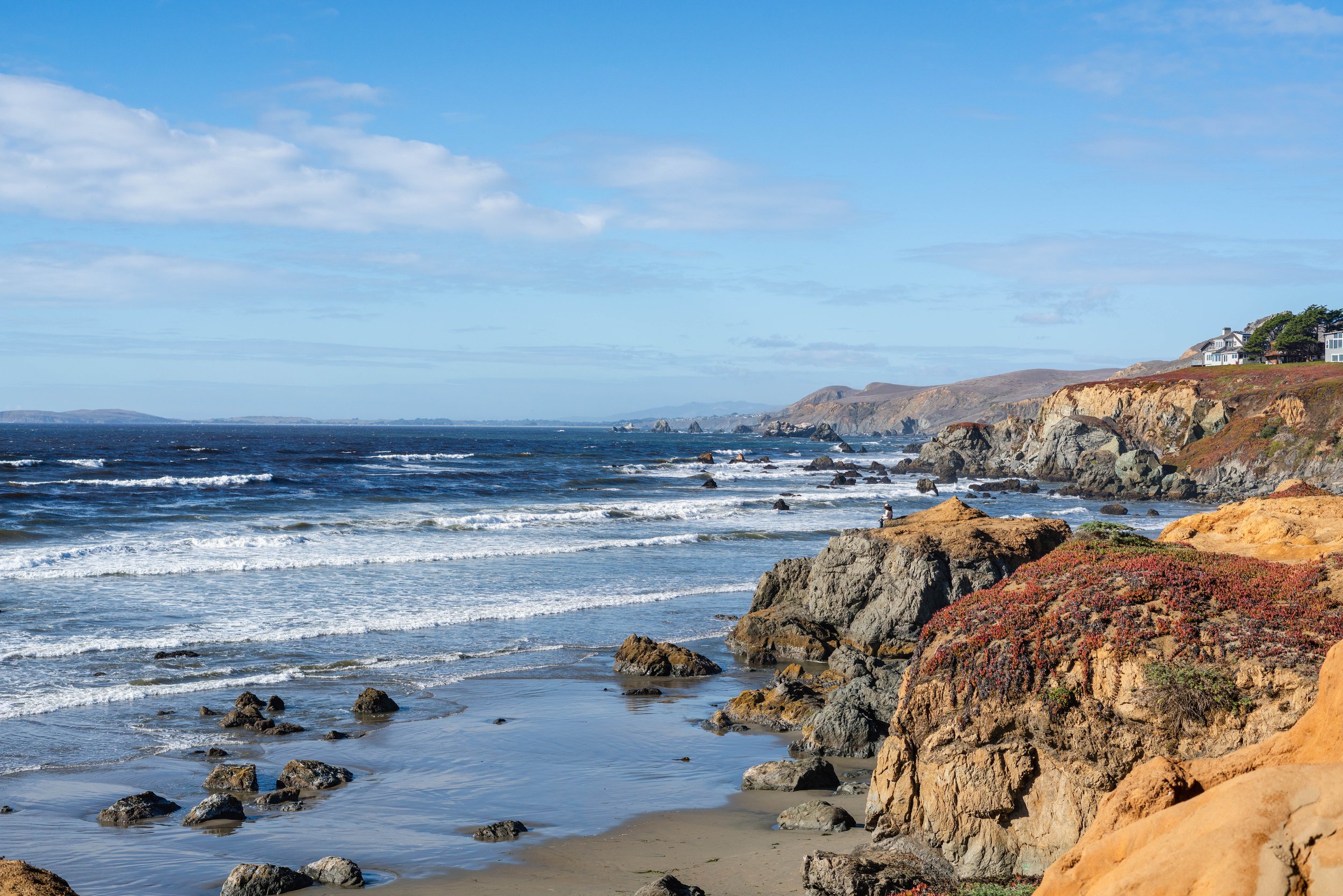 Coastal scene with rocky shoreline, waves, and a house on a hill.