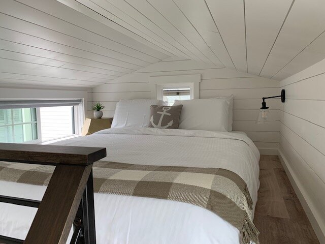 Cozy loft sleeping area in a tiny home with a queen bed, nautical decor, and a low vaulted ceiling.
