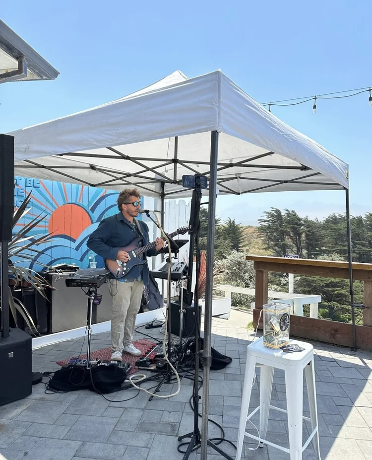 A man playing guitar and singing into a microphone under a white canopy, with a colorful mural behind him showing a sunrise or sunset over blue waves, outdoor setting with trees and sky.
