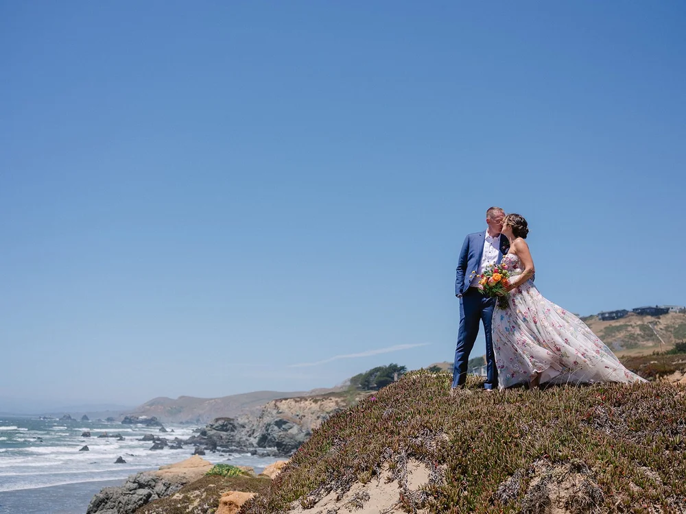 A bride and groom share a kiss on a grassy hill overlooking the ocean, with cliffs in the background under a clear blue sky.