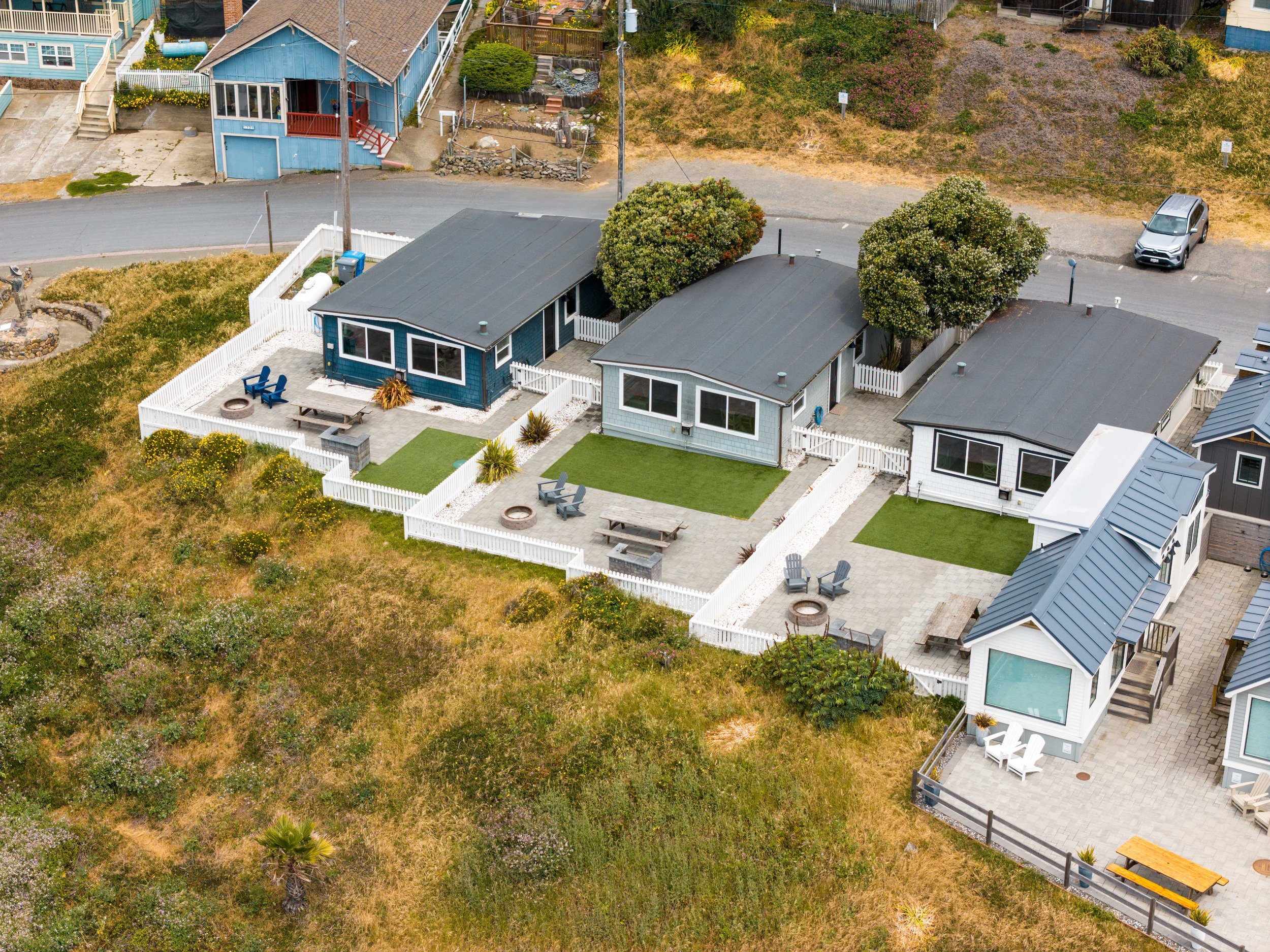 Aerial view of multiple coastal cottages at ブライトリビエラ, each with white picket fences, private gravel patios, fire pits, and artificial grass lawns, all overlooking the grassy coastal dunes.