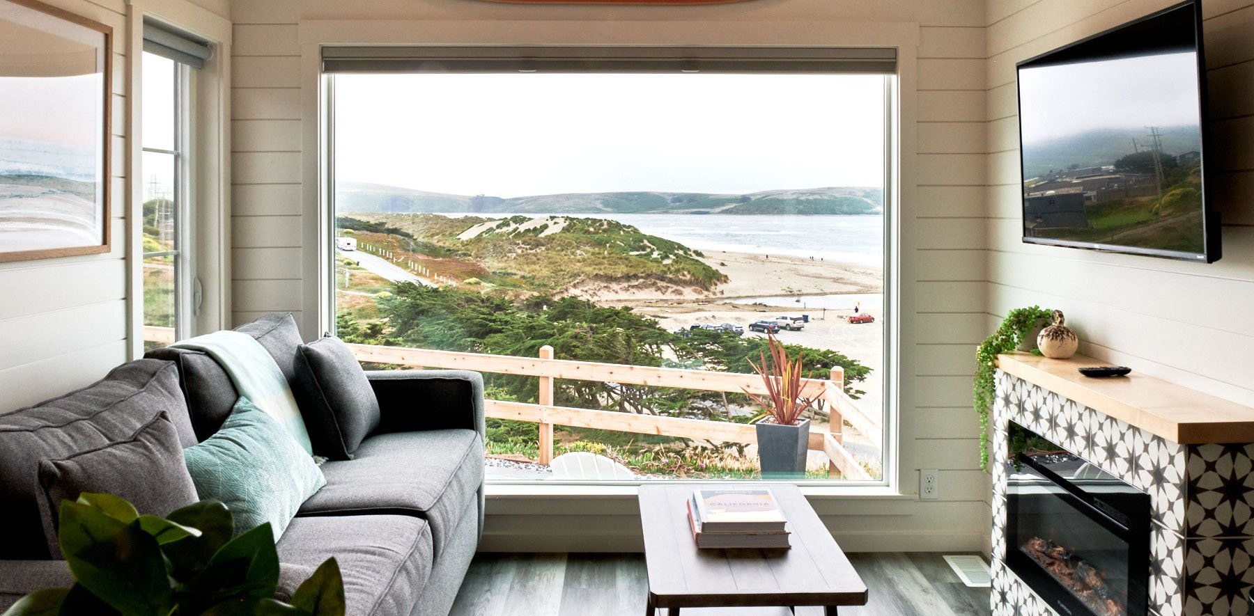 Living room with large window view of beach and dunes, gray sofa with pillows, wooden coffee table with books, and fireplace with decorative items and a mounted flat-screen TV.