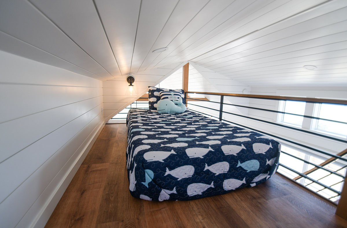A cozy loft bedroom in a ブライトリビエラ COHO cabin featuring a twin bed with a blue whale-patterned comforter, white shiplap walls, and a secure black metal railing overlooking the living area.