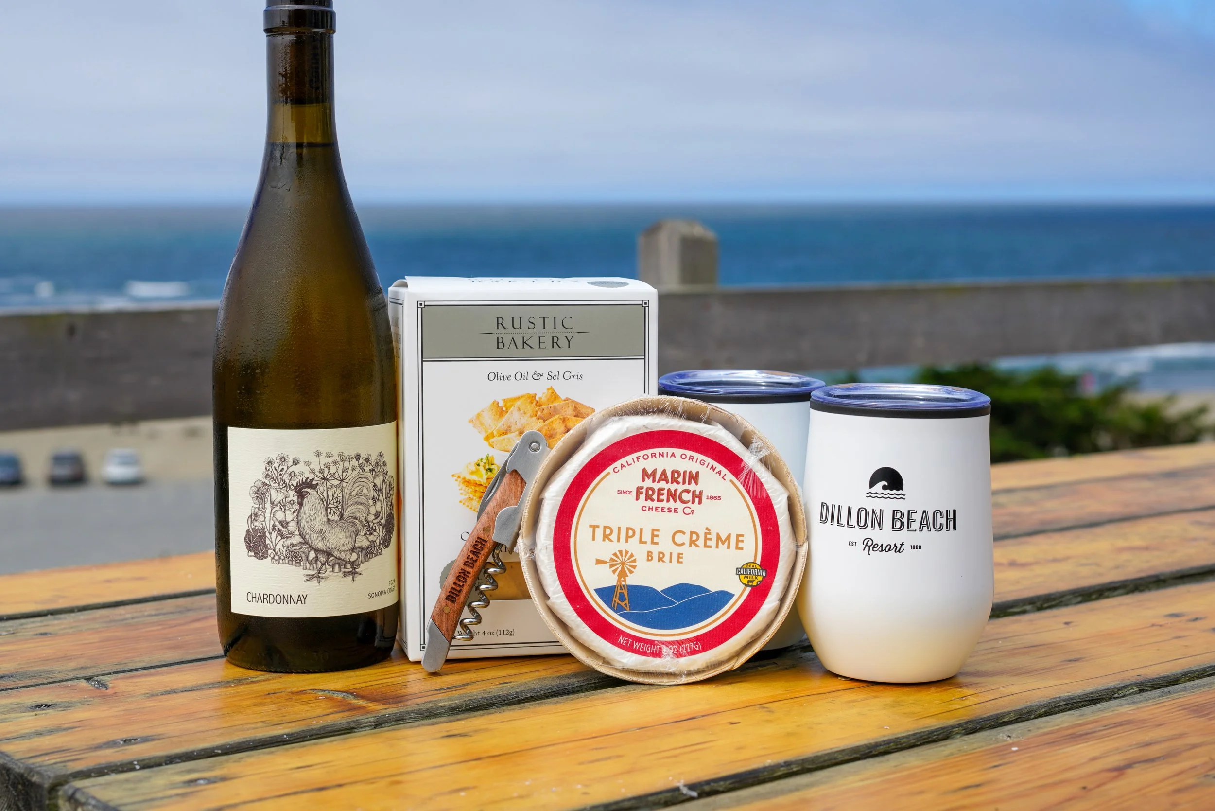 A bottle of Chardonnay wine, a box of Rustic Bakery olive oil and sea salt crackers, a package of フランス産 triple crème brie cheese, and two white cups, with a cloudy sky and ocean in the background, on a weathered wooden table.