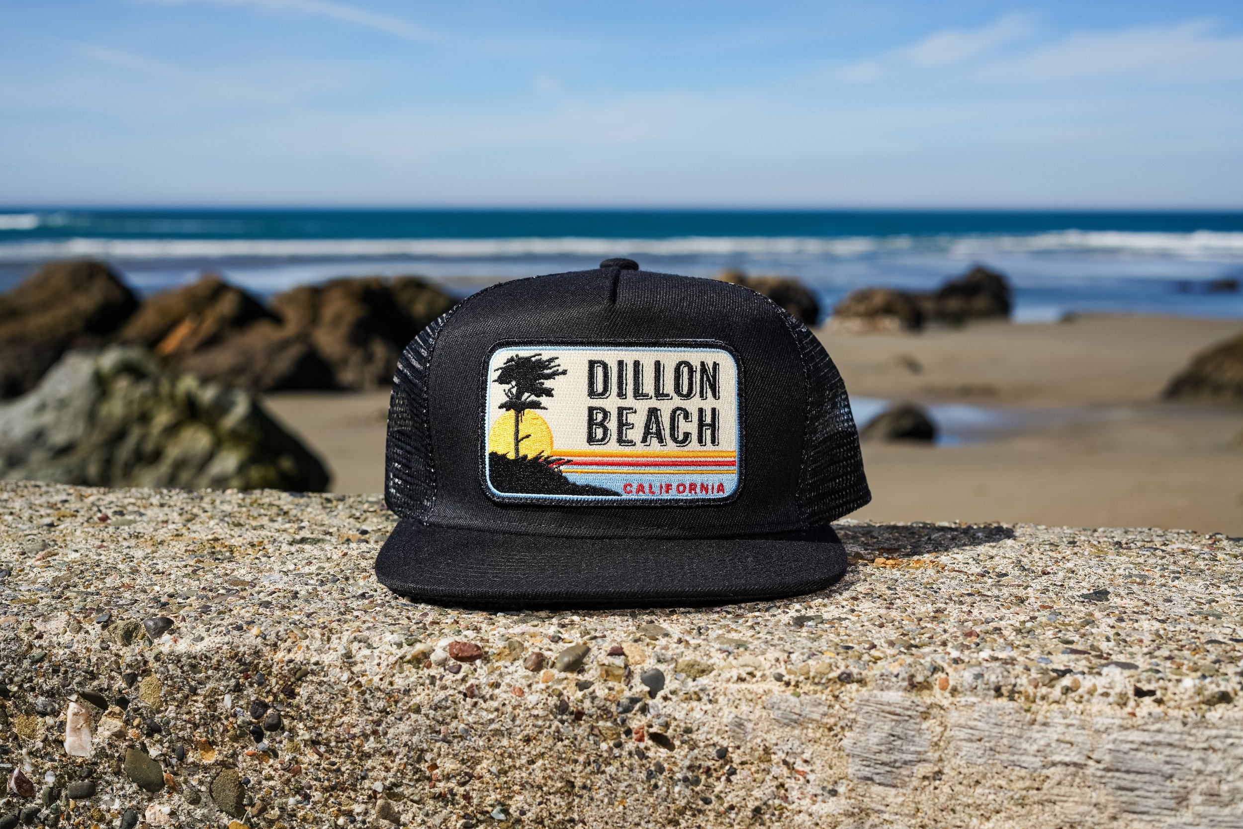 A black baseball cap with a ブライトリビエラパッチ on it, placed on a concrete surface at the beach with rocks and the ocean in the background.