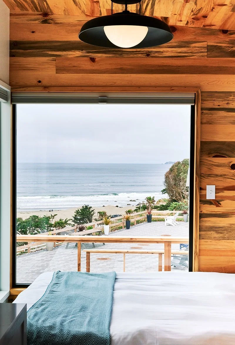 Interior of a modern Osprey cabin at ブライトリビエラ featuring a gray sofa with a yellow textured pillow, mid-century modern coffee table, and large floor-to-ceiling windows overlooking grassy dunes and the ocean.